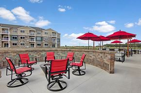 Sartell apartment for rent - A community patio here at Club at Heritage with cushioned seating and a fire pit area. Red chairs and umbrellas add a vibrant touch, while open views and surrounding buildings create a welcoming outdoor gathering space.