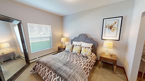 Sartell apartment for rent - A bedroom here at Club at Heritage featuring a cozy layout with a patterned bedspread, gray upholstered headboard, matching wood nightstands with lamps, and a large floor mirror adding brightness and depth.
