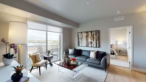 St. Paul apartment for rent - A stylish living room here at Collection at Highland Bridge featuring a modern sofa with accent pillows, a glass coffee table, a soft area rug, and a side chair positioned near sliding glass doors that open to a private balcony, with neutral walls, contemporary artwork, and warm natural light creating an inviting, comfortable atmosphere.