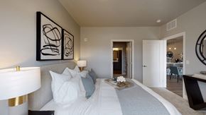St. Paul apartment for rent - A serene bedroom here at Collection at Highland Bridge featuring a neatly made bed with layered white and gray bedding, a padded headboard, matching bedside lamps, abstract wall art, soft carpet flooring, and an open doorway leading toward a bathroom and living area, creating a calm and polished retreat with a cohesive modern style.