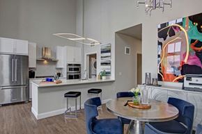Minneapolis apartment for rent - A stylish open kitchen and dining area here at Daymark Uptown featuring white cabinetry, a large island with bar seating, and stainless-steel appliances. A round dining table with upholstered chairs sits beneath modern lighting, while wood-style flooring, contemporary artwork, and a nearby hallway create a polished, open-concept layout.