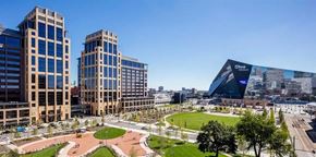 Minneapolis apartment for rent - A panoramic cityscape view here at Edition overlooking a landscaped urban park with walking paths, open green lawns, and surrounding modern high-rise buildings. The scene features a prominent glass-fronted stadium nearby, tree-lined streets, and expansive blue skies that highlight the vibrant downtown setting.
