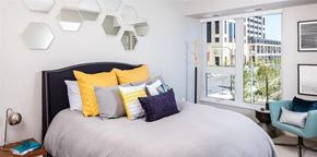 Minneapolis apartment for rent - A stylish bedroom here at Edition showcasing a modern bed with layered neutral and yellow accent pillows, a textured upholstered headboard, and decorative wall mirrors above. Large windows bring in natural light and offer city views, while coordinated furnishings and soft tones create a comfortable, polished retreat.