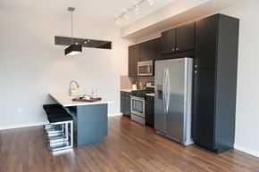Minneapolis apartment for rent - A modern kitchen here at Edition showcasing sleek dark cabinetry, a full suite of stainless steel appliances, and a large island with a smooth white countertop and integrated sink. Pendant and track lighting brighten the space, while wood-style flooring and clean lines create a contemporary, open-concept layout ideal for cooking and entertaining.