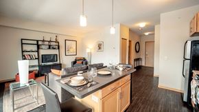 Hopkins apartment for rent - A modern, open kitchen here at Gallery Flats with light wood cabinets, dark countertops, and a center island set with bar seating and place settings. Pendant lighting hangs overhead, and the layout flows into a cozy living area with contemporary furniture, shelving, and soft accent lighting over dark wood flooring.