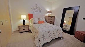 Hopkins apartment for rent - A cozy bedroom here at Gallery Flats with soft beige carpet, neutral walls, and a comfortable bed dressed in patterned bedding with accent pillows. Matching bedside tables with lamps provide warm lighting, while a tall dresser and a full-length mirror add function and style to the inviting space.