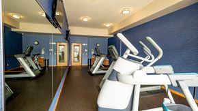 Hopkins apartment for rent - A modern fitness center here at Gallery Flats featuring cardio machines arranged along mirrored walls that reflect the space and lighting. Blue accent walls, dark rubber flooring, and ceiling-mounted lights create a clean, energized environment, while a wall-mounted television and glass doors add convenience and visibility.