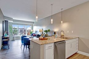 Minneapolis apartment for rent - A bright, open kitchen here at Gatsby featuring white cabinets, stone countertops, and a large island with an integrated sink and seating space. Pendant lights hang overhead, and the layout opens to a living and dining area with wood-style flooring and large windows that bring in natural light and views of nearby buildings.