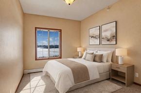 St. Cloud apartment for rent - A cozy bedroom here at Grand River Estates featuring soft neutral walls, plush carpet flooring, and a comfortable bed with layered white bedding and accent pillows. The room includes matching nightstands with lamps, framed wall art above the headboard, and a window that brings in natural light and outdoor views, creating a calm and inviting sleeping space.
