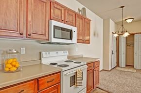St. Cloud apartment for rent - A warm kitchen here at Grand River Estates showcasing rich wood cabinetry, generous countertop space, and a clean, functional layout designed for everyday cooking. The space features ample storage, coordinated appliances, neutral countertops, and an open connection to the dining area with soft lighting and carpeted flooring extending beyond the kitchen.