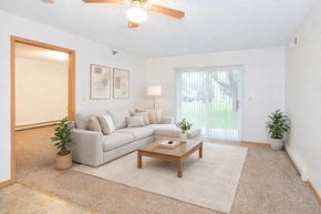 St. Cloud apartment for rent - A bright living room here at Heritage Park East featuring a spacious open layout with plush carpeting and a neutral color palette. The room includes a light-toned sectional sofa with accent pillows, a wooden coffee table atop a soft area rug, a ceiling fan with overhead lighting, and sliding glass doors that bring in natural light and views of green outdoor space.