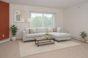 St. Cloud apartment for rent - A bright living room here at Heritage Park East showcasing a modern, open layout with soft carpeting and a clean neutral color palette. The space features a light gray sectional sofa with textured accent pillows, a wooden coffee table with a black metal frame, potted greenery, abstract wall art, and a large window with vertical blinds that brings in natural light and outdoor views.