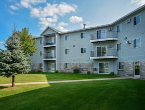 St. Cloud apartment for rent - A well-maintained apartment community exterior here at Heritage Park East featuring a three-story residential building with light gray siding, private balconies, and large windows overlooking a lush green courtyard. The landscaped lawn includes mature trees, trimmed shrubs, and a paved walking path, creating an open and inviting outdoor setting for residents.