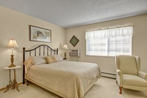 Saint Cloud apartment for rent - A comfortable bedroom here at Heritage Park featuring a neatly made bed with soft neutral bedding, matching bedside tables with warm accent lamps, and tasteful wall art that adds character. Natural light filters through a wide window with blinds, complementing the plush carpet and creating a calm, inviting retreat.