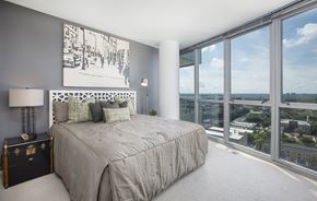 Minneapolis apartment for rent - A sunlit bedroom here at LPM Apartments featuring a comfortable bed dressed in soft neutral bedding and layered accent pillows, set against a gray accent wall with modern artwork, while expansive floor-to-ceiling windows wrap the space with sweeping city views, filling the room with natural light and creating an airy, serene retreat.