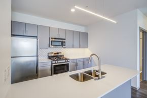 Minneapolis apartment for rent - A sleek modern kitchen here at LPM Apartments featuring smooth light countertops with an integrated sink and curved faucet, gray flat-panel cabinetry, stainless-steel appliances, and a geometric tile backsplash, accented by linear pendant lighting and an open island layout that creates a clean, bright, and contemporary cooking and entertaining space.