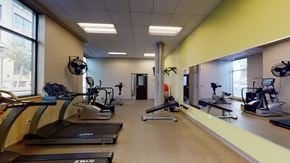 St. Paul apartment for rent - A modern fitness center here at Lofts at Farmers Market featuring treadmills and cardio machines lined along large windows, mirrored walls that reflect the workout space, a weight bench and exercise equipment for strength training, neutral flooring, light-colored walls, ceiling lighting, and an open layout designed for convenient and comfortable workouts.