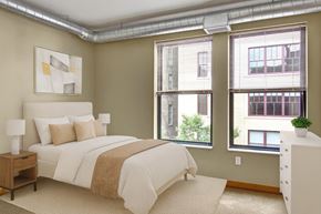 St. Paul apartment for rent - A serene bedroom here at Lofts at Farmers Market featuring a plush bed with neutral bedding and accent pillows, exposed ductwork along the ceiling, and large windows with blinds that bring in natural light, complemented by soft carpeting, a minimalist nightstand with lamp, and clean modern finishes for a calm, loft-style retreat.