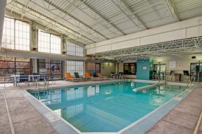 Minneapolis apartment for rent - A spacious indoor pool here at Marbella on Dean featuring a large rectangular swimming pool with clear blue water, stainless steel handrails, surrounding concrete deck, and lounge seating with bright accent chairs, set beneath a high industrial-style ceiling with exposed beams and large windows that bring in natural light.