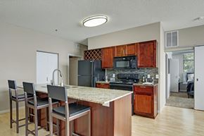 Minneapolis apartment for rent - A warm open kitchen here at Marbella on Dean featuring rich wood cabinetry, a spacious island with granite-style countertops and bar seating, a tile backsplash, and a full appliance suite, with light wood flooring, a built-in wine rack, and an adjacent hallway leading toward the bedroom area creating a functional and inviting layout.