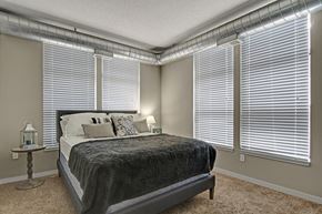 Minneapolis apartment for rent - A cozy sunlit bedroom here at Marbella on Dean featuring a plush bed with layered neutral bedding, soft carpeted flooring, and expansive corner windows with blinds that fill the space with natural light, complemented by exposed ductwork along the ceiling, light-toned walls, and a warm, inviting layout ideal for rest and relaxation.