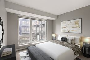 Minneapolis apartment for rent - Modern bedroom here at Maverick with a white bed, patterned throw, dark bench, large window, and wall art in a neutral-toned room.
