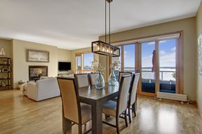 Spring Park apartment for rent - A bright open dining and living area here at The Mist featuring light wood-style flooring, a rectangular dining table with upholstered chairs, and a modern pendant light overhead. The space flows into a cozy living room with a fireplace and seating, while large glass doors and windows showcase scenic water views and lead to a private balcony.