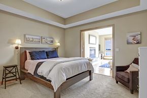 Spring Park apartment for rent - A serene bedroom here at The Mist featuring a spacious layout with a wood-framed bed dressed in soft neutral bedding, matching wall-mounted lamps, and framed coastal artwork above the headboard. Plush carpeting adds comfort underfoot, while an open doorway leads to a bright adjoining living area with large windows that bring in abundant natural light and scenic views.