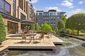 Spring Park apartment for rent - A scenic outdoor patio here at The Mist featuring café-style tables and chairs arranged on a spacious stone terrace overlooking a tranquil water feature. Lush landscaping, manicured trees, and surrounding apartment buildings frame the setting, creating a peaceful space for relaxing outdoors while enjoying fresh air and sunny views.