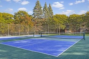 Plymouth apartment for rent - A scenic outdoor tennis court here at Plymouth Square featuring blue and green playing surfaces, a central net, and surrounding fencing, set against a backdrop of mature trees and nearby apartment buildings. The open layout and clear markings create an inviting space for residents to enjoy casual matches or practice in a peaceful, park-like setting.