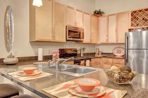Ramsey apartment for rent - A contemporary kitchen here at Residence at the COR showcasing light wood cabinetry, sleek countertops with an integrated double sink, stainless-style appliances, and a built-in microwave, complemented by a breakfast bar set with place settings, decorative accents, and warm pendant lighting that creates a functional and inviting cooking space.