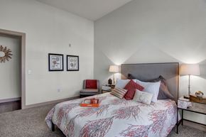 Ramsey apartment for rent - A cozy bedroom here at Residence at the COR showcasing a comfortable bed with layered neutral and warm accent pillows, a patterned comforter, and matching bedside tables with soft lighting, complemented by plush carpeting, simple wall art, and a small seating area that creates a calm, welcoming space designed for everyday comfort and relaxation.