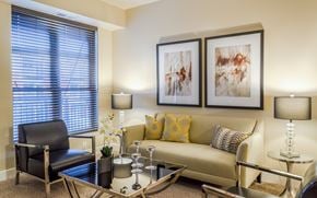 St. Louis Park apartment for rent - A warm and inviting living room here at Siena featuring a comfortable sofa accented with patterned throw pillows, a sleek glass coffee table, and modern table lamps that provide soft lighting, complemented by contemporary wall art, a cozy accent chair, and a large window with blinds that brings in natural light and creates a relaxed, welcoming atmosphere.