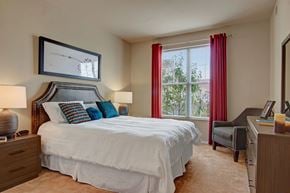 Plymouth apartment for rent - A cozy bedroom here at Stoneleigh at the Reserve featuring a comfortable bed with crisp white bedding and colorful accent pillows, complemented by matching nightstands with lamps, soft carpeted flooring, and neutral walls, while a large window with curtains brings in natural light and creates a calm, inviting retreat for rest and relaxation.