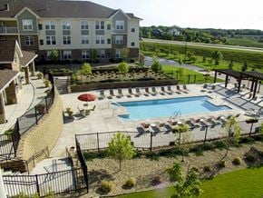 Plymouth apartment for rent - A sunlit outdoor pool here at Stoneleigh at the Reserve featuring a large rectangular swimming pool surrounded by a spacious concrete deck, rows of lounge chairs with small tables, and a shaded seating area with umbrellas, all enclosed by black metal fencing and framed by landscaped greenery, walking paths, and neighboring apartment buildings under open blue skies.