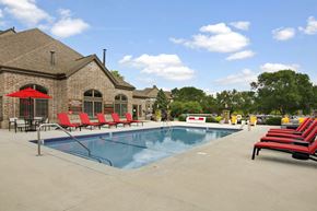 Minneapolis apartment for rent - A resort-style outdoor swimming pool here at The Fairway featuring clear blue water with metal handrails, a wide concrete pool deck, and rows of red cushioned lounge chairs arranged for relaxation, alongside a brick clubhouse building with arched windows, shaded seating areas, mature trees, and open skies creating a welcoming community gathering space.
