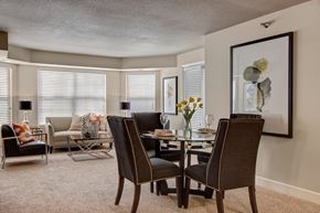 Minneapolis apartment for rent - A welcoming living and dining area here at The Fairway showcasing a glass-top dining table with upholstered chairs, plush carpet flooring, and a comfortable seating area with a sofa, accent chair, and metallic coffee table, complemented by framed artwork, table lamps, and large windows with blinds that bring in soft natural light.
