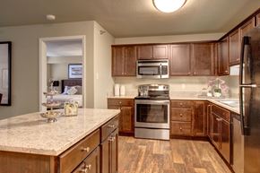 Minneapolis apartment for rent - A classic kitchen here at The Fairway featuring rich wood cabinetry, speckled countertops, and a spacious island with storage, complemented by a built-in microwave, oven, and refrigerator, warm wood-style flooring, and soft overhead lighting that creates a functional and inviting space connected to the adjacent bedroom.