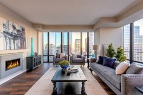 Minneapolis apartment for rent - A stylish living room here at The Nic on Fifth showcasing rich wood-style flooring, a contemporary sofa and matching armchairs with accent pillows, and a dark wood coffee table centered on a light area rug, complemented by a sleek fireplace, modern wall art, greenery accents, and expansive floor-to-ceiling windows framing stunning city skyline views.