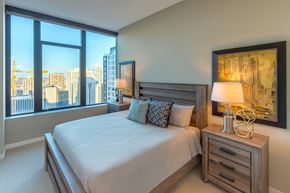 Minneapolis apartment for rent - A sunlit bedroom here at The Nic on Fifth featuring a modern wood-frame bed dressed in soft neutral bedding, matching nightstands with sculptural lamps, and contemporary artwork, while expansive floor-to-ceiling windows frame sweeping city views, filling the space with natural light and creating a refined, comfortable retreat.
