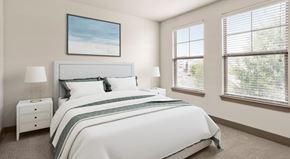 Charlotte apartment for rent - A bright bedroom here at CityPark View featuring a neatly made bed with crisp white bedding, a padded headboard, and matching bedside tables with lamps. Soft carpet flooring and neutral walls create a calm feel, while large windows with blinds allow natural light and offer views of surrounding greenery.