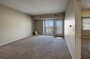 Williston apartment for rent - A spacious, unfurnished living room here at Dakota Commons featuring soft neutral walls, plush carpet flooring, and a sliding glass door with an adjacent exterior door that opens to a private balcony, allowing natural light to fill the open layout and highlight the room’s generous size.