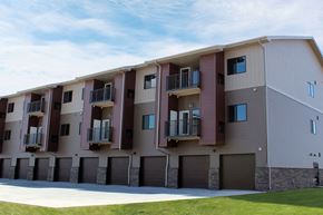 Williston apartment for rent - A modern apartment building exterior here at Dakota Commons featuring a three-story structure with private balconies, ground-level garages, mixed neutral siding with brick accents, and a well-maintained lawn in the foreground beneath a bright blue sky.