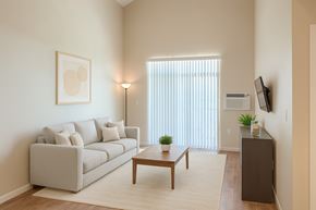 Williston apartment for rent - A bright, modern living room here at Dakota Ridge featuring high ceilings, a light neutral sofa with coordinating pillows, a wooden coffee table on a soft area rug, warm wood-style flooring, vertical blinds allowing natural light, a wall-mounted TV, and minimal decor creating an open, inviting atmosphere.