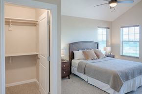 Williston apartment for rent - A bright, comfortable bedroom here at Dakota Ridge featuring a neatly made bed with neutral bedding, matching nightstands with lamps, soft carpeted flooring, a ceiling fan with light, two sunlit windows with blinds, and an open walk-in closet with built-in shelving for convenient storage.