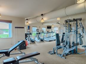 Williston apartment for rent - A spacious, well-lit fitness center here at Dakota Ridge featuring treadmills lined along large windows, a multifunction weight machine, a padded workout bench, neutral carpet flooring, exposed ceiling ductwork, and warm pendant lighting that creates a clean, motivating environment for residents to exercise comfortably.