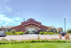 Williston apartment for rent - A welcoming exterior view here at Dakota Ridge featuring the brick clubhouse entrance with a peaked roofline, glass double doors, landscaped greenery, colorful planters, and a wide parking area set against a bright blue sky and well-maintained lawns.