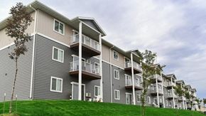 Watford City apartment for rent - A modern apartment building exterior here at Pheasant Ridge showcasing multi-story residences with gray and beige siding, private balconies, white railings, evenly spaced windows, and a well-maintained green lawn with young trees under a lightly overcast sky.
