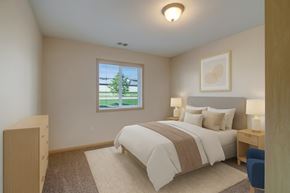 Watford City apartment for rent - A cozy bedroom here at Pheasant Ridge featuring soft neutral walls, a queen-sized bed with white bedding and a taupe accent throw, matching bedside tables with modern lamps, a light area rug over plush carpet, minimal wall art above the headboard, and a window that brings in natural light and views of green landscaping.