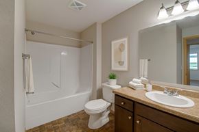 Watford City apartment for rent - A clean bathroom here at Pheasant Ridge featuring a white tub and shower combination with smooth surround walls, a single-sink vanity with dark wood cabinetry and a neutral countertop, a large wall-mounted mirror with bright overhead lighting, folded white towels, simple wall art, and durable flooring in warm earth tones.